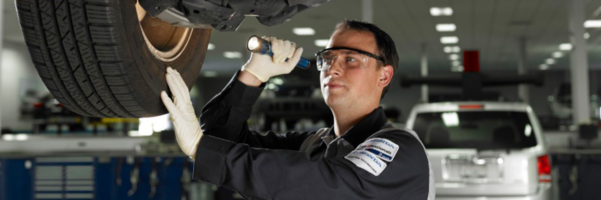 Honda service technician inspecting underneath car on lift with flashlight.