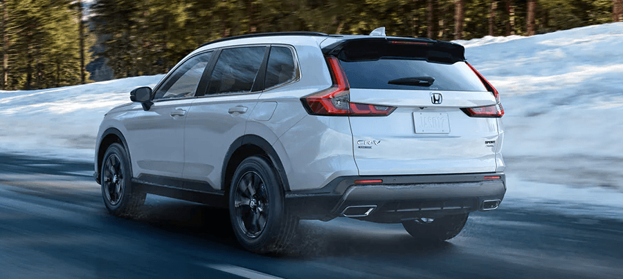 Rear driver-side view of the 2023 Honda CR-V Sport Touring Hybrid in Platinum White Pearl, driving on a mountain road with snow and trees in the background.
